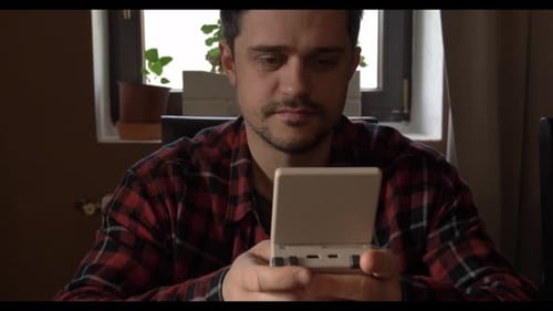 Video footage of a middle-aged man in a plaid shirt playing a game on a portable retro console, view