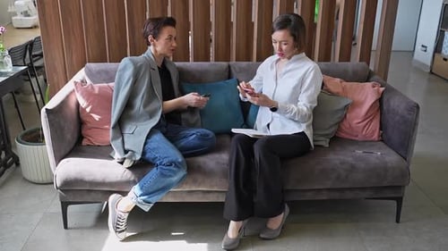 Two Women Using Laptop and Mobile Phone Together on Sofa with Wooden Wall in Office Collaboration