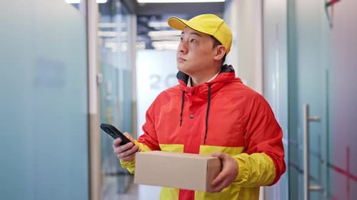 Confident Male Courier Using App for Delivery on Phone in Corridor with Doors