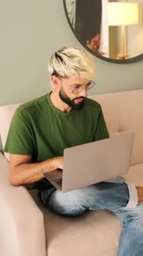 Young blond man with a beard works on a laptop while sitting in a cozy living room