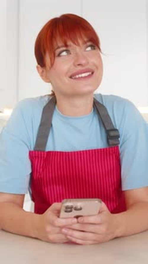 Young Woman in Apron at Bar Uses Smartphone Sees Good News Smiles Celebrates Win with Happy Bounce