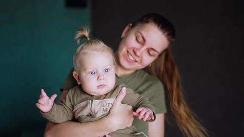 Mother is holding her little child. Cute baby with blond hair looks ahead with surprise.