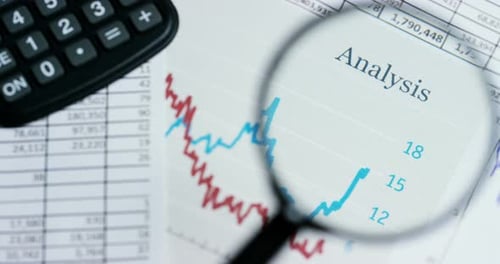 A hand with a magnifying glass passes over a financial newspaper and financial bag with a business