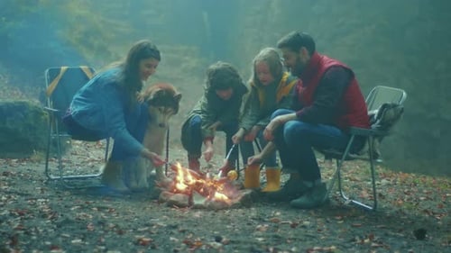 Happy Family with Dog Sit Around a Campfire on Autumn Evening in the Woods Frying Sausages Over a