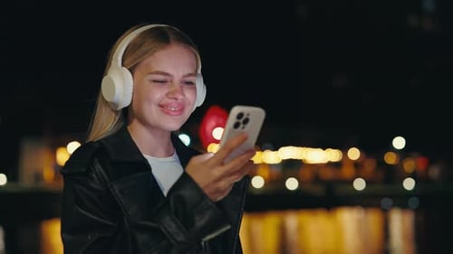 Pretty Girl Using Phone Wearing White Headphones Standing Outside at the Night City