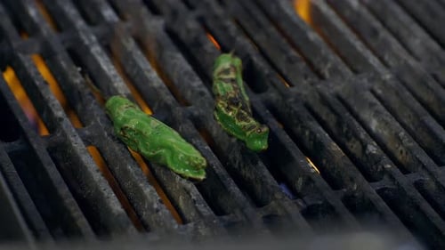 Shishito peppers on a grill in a kitchen are turned over with silver tongs