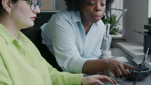 Two Multiethnic Female Office Employees Talking and Using Laptop