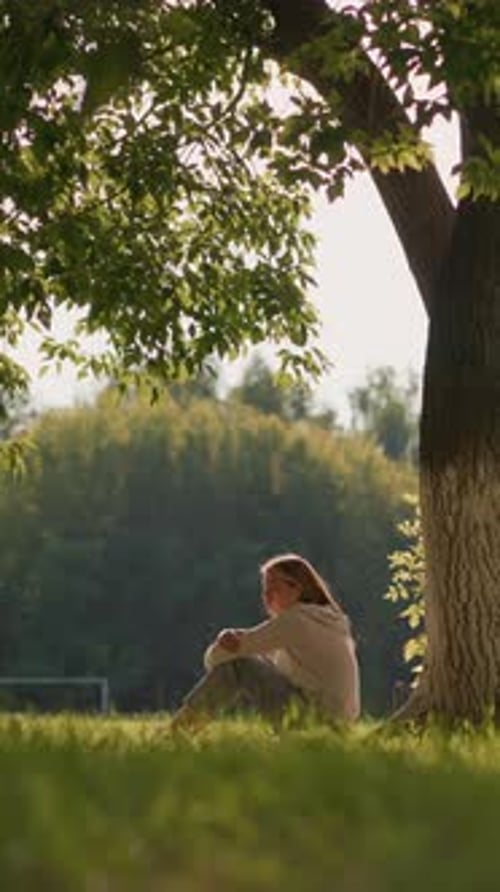 Woman Sitting Under Tree in Sunlit Park Surrounded By Greenery and Gentle Wind
