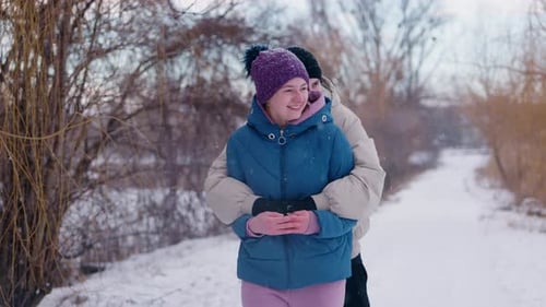 Junges Paar genießt eine verspielte Umarmung auf einem verschneiten Pfad, umgeben von Winterbäumen, die warm lächeln
