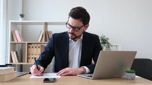Thoughtful Businessman Taking Notes at Office Desk with Laptop and Documents
