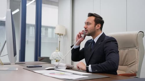 Businessman in a suit talks on the phone and points at his computer screen in a modern office