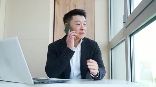 young asian male businessman talking on cellphone sitting at office desk in business formal suit