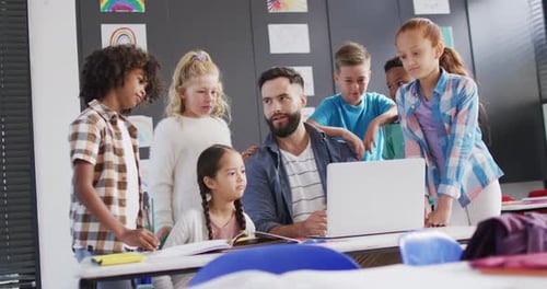 Diverse male teacher and happy schoolchildren using laptop in school classroom