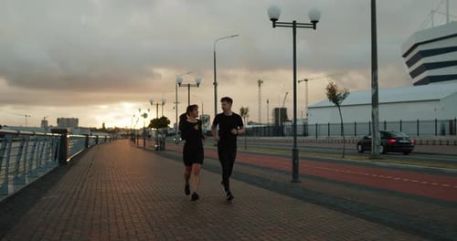 Young Man and Woman Running Along the Embankment Near the Stadium at Sunset