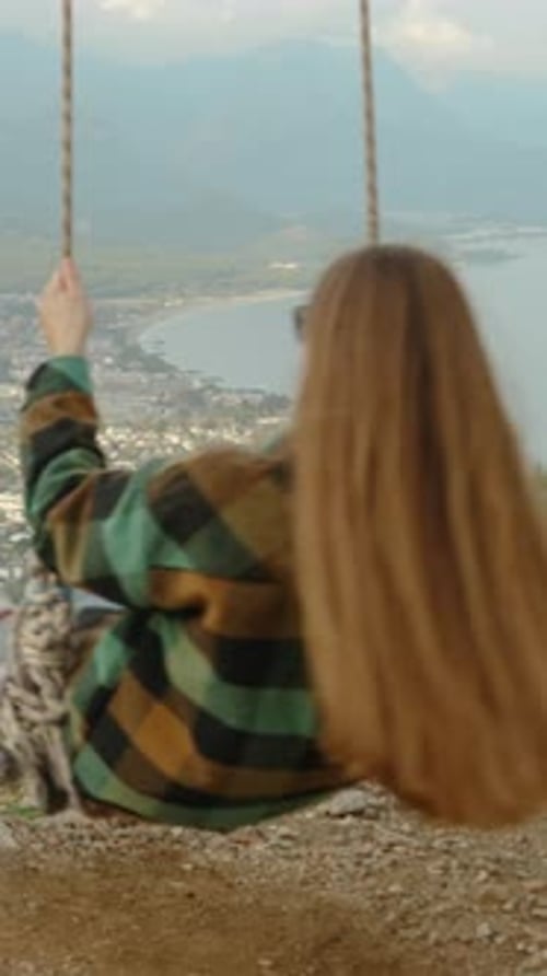 Vertical video. A panoramic view circles around the young woman swinging on swings over the cliff.