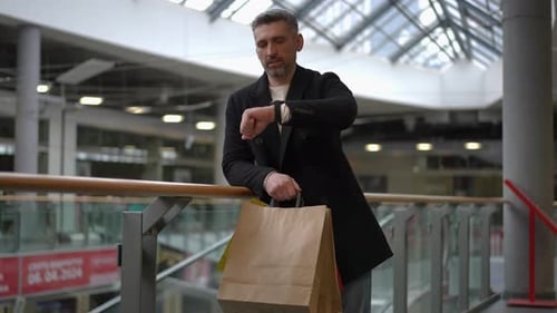 Man Checking Time While Holding Shopping Bags in a Modern Mall