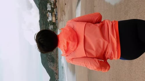 Female in orange jacket running along seashore on sandy beach. Vertical video