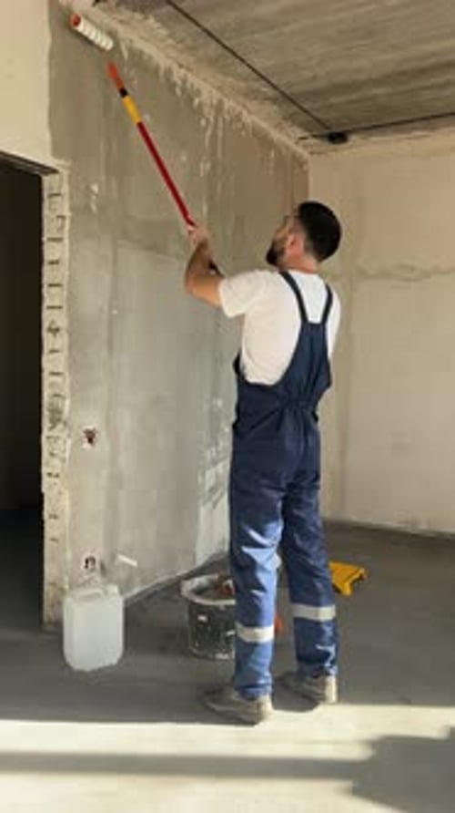 A Bearded Man in a Work Uniform is Priming the Walls After Applying Gypsum Plaster