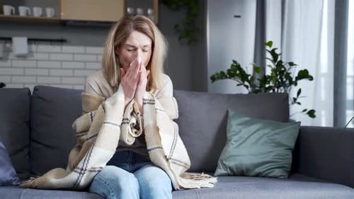 Femme blonde malade avec une tasse de thé chaud assise sur le canapé sous la couverture chaude et a une tête sévère