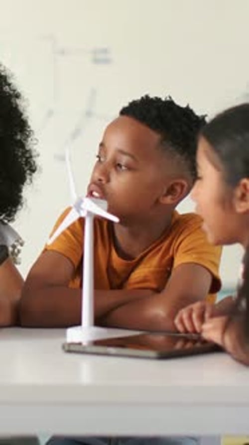 Vertical video: Leaning boy in orange tee checking wind turbine in STEM lesson, tablet, whiteboard