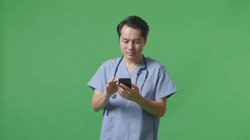 Asian Male Doctor With Stethoscope Using Smartphone While Standing On Green Screen Background