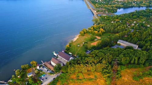 Hotel located on the river bank with few piers and boats. Green waterfront and dam at backdrop.