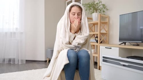 Jeune femme ayant froid à la maison se réchauffant sous une couverture à côté d'un radiateur électrique.