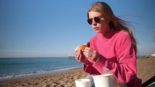 young woman sitting on the beach in cool weather and eating sandwich