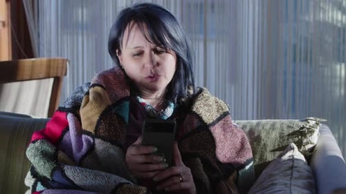 Woman Wrapped in Blanket Looking at Smartphone on Couch