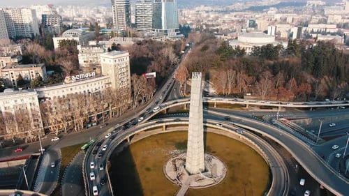Cars Navigating a Roundabout Featuring a Tall Slender Monument at Its Center Surrounding Buildings