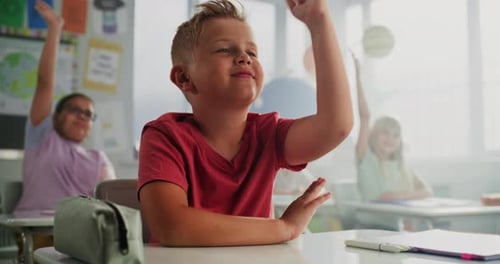 Elementary School Boy Sitting at Desk Raising Hand and Answering a Teacher's Question