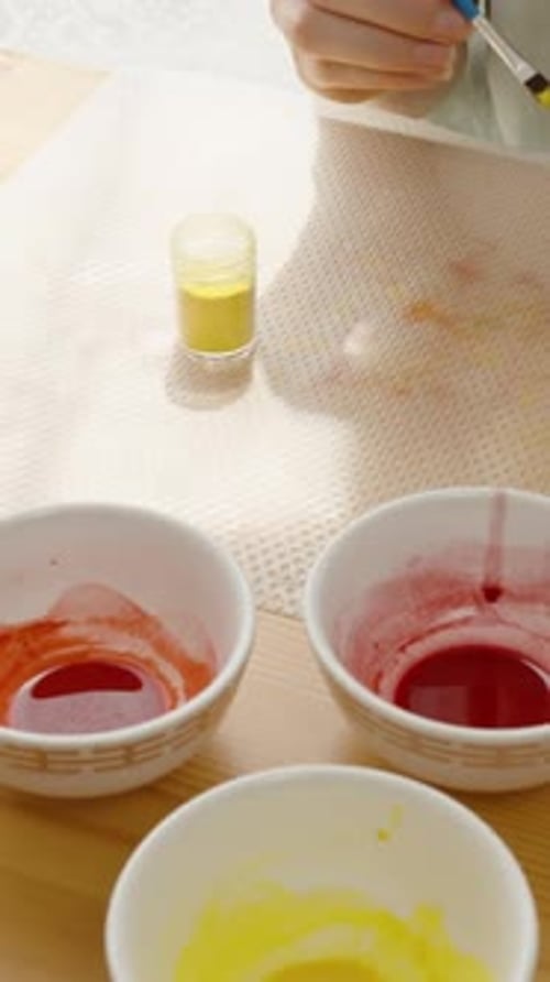 Vertical video. Woman uses a brush to paint a marbled pink and yellow Easter egg, with a small jar
