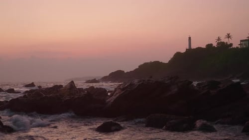 Lighthouse Stands on a Hill As Sunset Paints Sky in Shades of Pink Purple Over Ocean Waves Crash