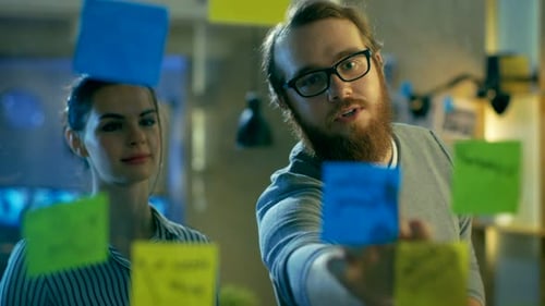 Male and Female Designers Plan Their Next Project on a Glass Wall with Sticky Notes Outline. They'r
