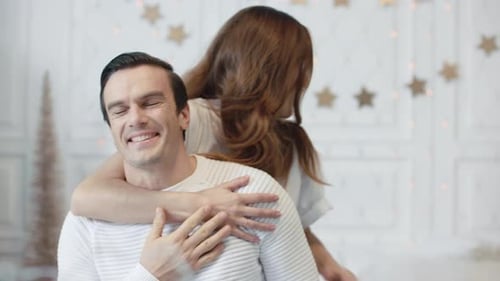 Smiling Wife Asking Husband Close Eyes In Living Room. Happy Woman Presenting Christmas Gift To B...