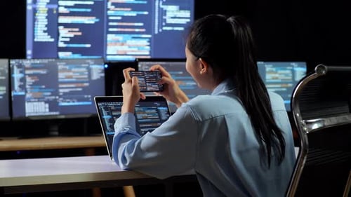 Back View Of Female Programmer Looking At Database On Smartphone While Writing Code By A Laptop