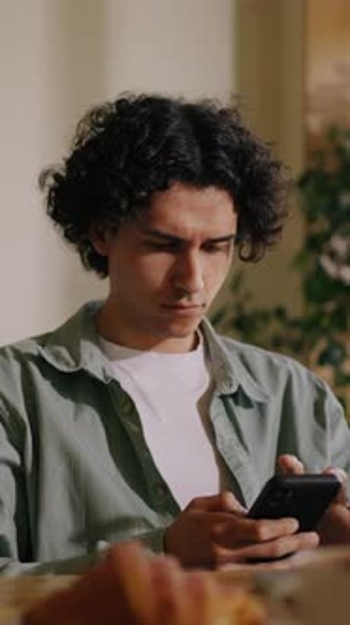 Thoughtful Young Man with Curly Hair Sitting Indoors Concentrating on Smartphone Screen While Typing
