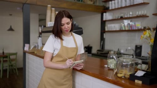 Portrait of a Brunette Waitress Girl in a Yellow Apron Who Makes Notes in Her Notebook Near the Cash
