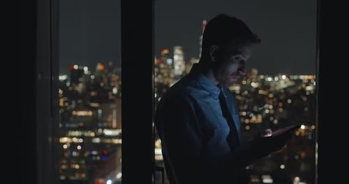 Businessman texting. Man standing near the windows at night urban background. Holding smartphone lig
