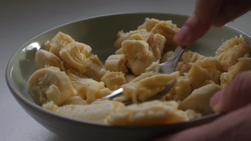 Person mashing chopped banana in small bowl with a metal fork on benchtop