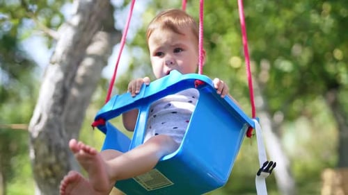 Sweet barefoot baby sits on a swing. Little kid having fun outdoors in the garden on warm sunny day.
