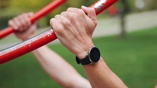 Focused young athletic man with smartwatch doing pull-ups at red sports facility in park