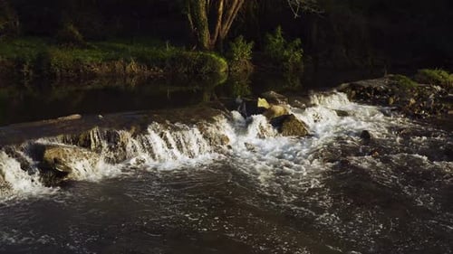 Water flowing down over a natural dam creating some green and white foam and water rapids. Sun shinn