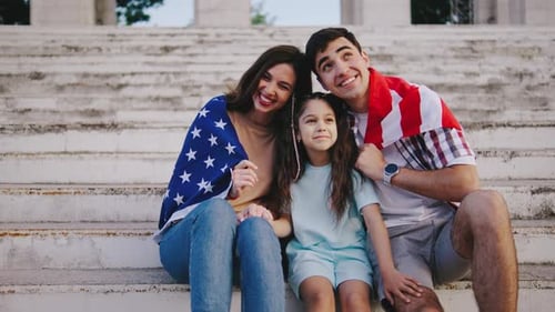 A Young Family with a Child Girl is Sitting on the Steps and Wrapped in the Flag of the United
