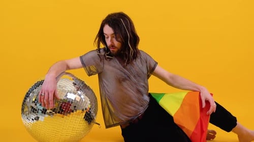 Gay Man Dressed in Mesh Tshirt Sits on Yellow Background with a Multicolored Flag and Silver Ball