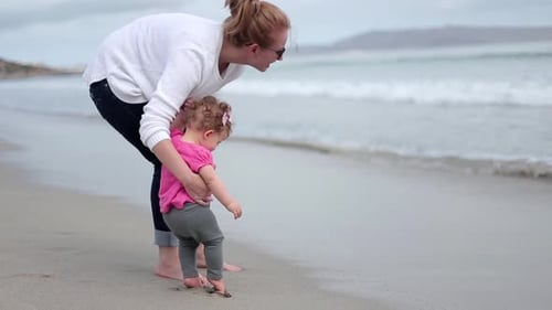 A mother holds her toddler carefully as she plays on the beach with the waves rolling in