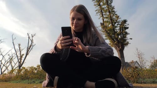 Young Attractive Blonde Girl Sitting Outdoors in Park Using Phone