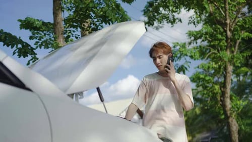 Young asian man having trouble with his broken car, opening hood and calling for help on cell phone