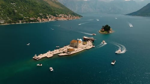 Cinematic aerial drone view of Our Lady of the Rocks island with its stone church, tourists, and sur