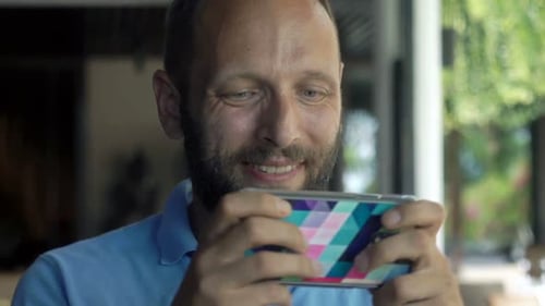 Happy, Young Man Playing Game on Smartphone Sitting in Cafe 30s